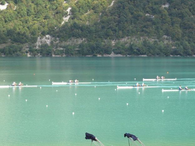 Gruppe von Menschen beim Ruderwettbewerb auf einem Gewässer mit Bäumen und einem Hügel im Hintergrund, Kleidung über Tribünen am Wasserrand gehängt.