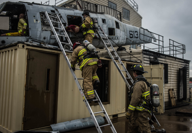 Feuerwehrleute in Uniform und Helmen arbeiten an einem Hubschrauber auf einem Gebäudedach, einer hält einen Sauerstoffzylinder, mit Leitern, einem Container, Bäumen und einem blauen Himmel im Hintergrund.