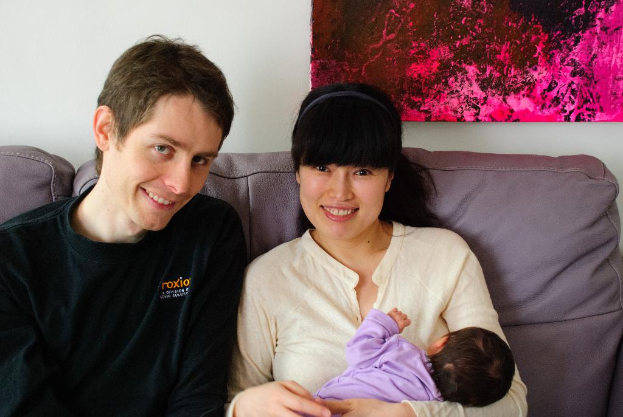 Eine Frau in einem wei├čen Hemd h├Ąlt ein Baby und l├Ąchelt, waehrend sie auf einem Sofa sitzt neben einem Mann in einem schwarzen T-Shirt, der ebenfalls l├Ąchelt, mit einem rosafarbenen Foto Rahmen an der wei├čen Wand dahinter.