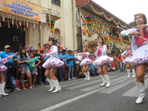 Eine Gruppe von Frauen in pinken und weißen Outfits, die auf der Straße tanzen, umgeben von einer Menge Menschen, die stehen und zusehen, mit Gebäuden, Bäumen und einem klaren blauen Himmel im Hintergrund.