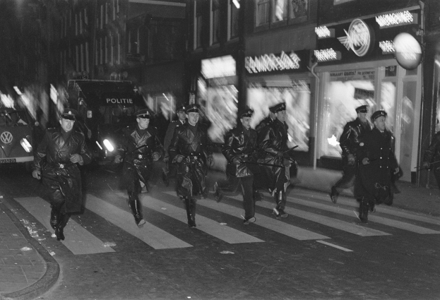 Schwarzes und weißes Bild von Polizisten in Uniform, die nachts über eine Straße gehen, einige halten Waffen, mit Fahrzeugen und Gebäuden im Hintergrund.