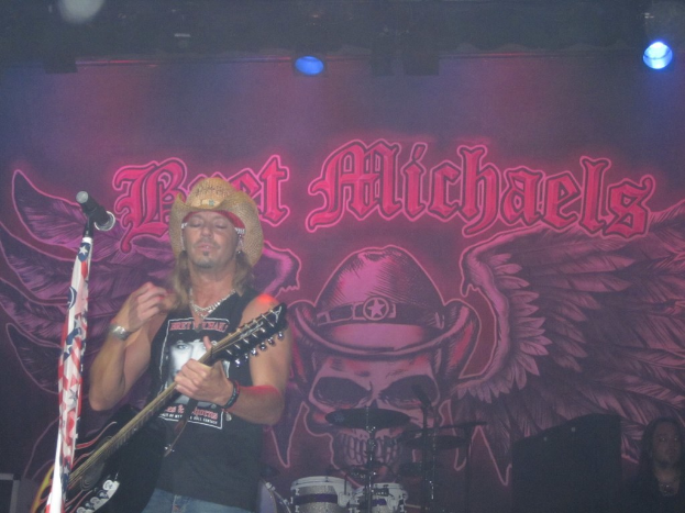 Eine Musikkonzertsituation mit Scheinwerfern oben, einem Banner im Hintergrund, einer Person mit Gitarre links mit Mikrofon davor, einer Person in der rechten unteren Ecke und Gegenständen, die wie Drogen aussehen, in der Mitte unten.