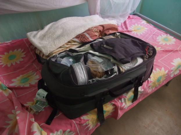 Ein Bett mit einer bunten Bettdecke, eine offene Gepäcktasche mit Kleidung und Tags mit Barcodes darauf, vor einer Wand in einem Raum.