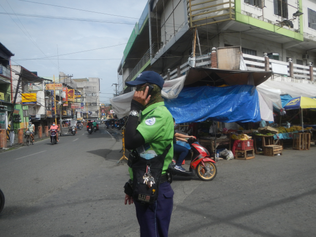 Ein Polizist in grüner und blauer Uniform und blauer Mütze steht an der Straße und spricht in ein Handy, während Fahrzeuge auf der Straße fahren, Stände mit verschiedenen Gegenständen rechts von ihm und ein bewölkter Himmel mit einem Gebäude mit Fenstern im Hintergrund.