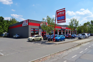 Ein Autohaus mit mehreren parkenden Fahrzeugen vorne, umgeben von Gebäuden, Bäumen, Strommasten, Schildern und einer Straße unter einem bewölkten Himmel.