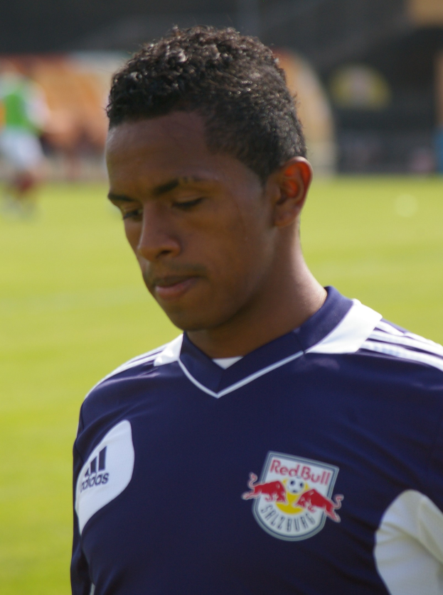 Ein junger Mann in einem blauen T-Shirt steht auf einem Fußballfeld, er wirkt wie ein Flügelspieler mit kurzen dunklen Haaren und einem entschlossenen Gesichtsausdruck vor einem unscharfen Hintergrund.