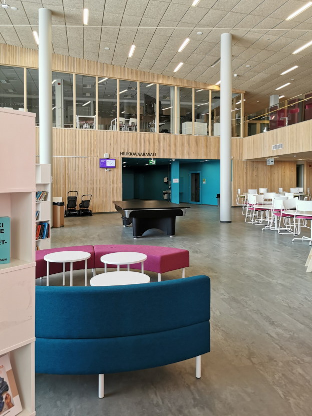 Großer Raum mit mehreren Tischen, Stühlen, Sofas, Hockern, Bücherregalen, Säulen, Deckenleuchten und einem wandmontierten Fernseher, der an ein Schulbibliotheksinnere erinnert.