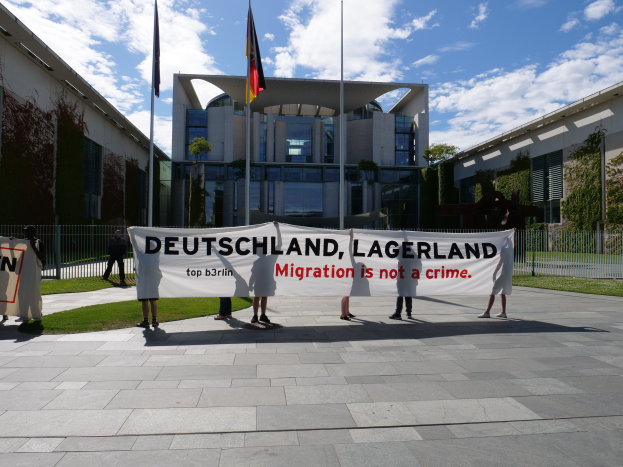 Eine Gruppe von Menschen hält ein Banner mit der Aufschrift "Deutschland, Lagerland Migration ist kein Verbrechen" vor einem Gebäude mit Fenstern, umgeben von Gras, einem Metallzaun, Pflanzen, Bäumen und Fahnen mit Stangen, unter einem bewölkten Himmel.