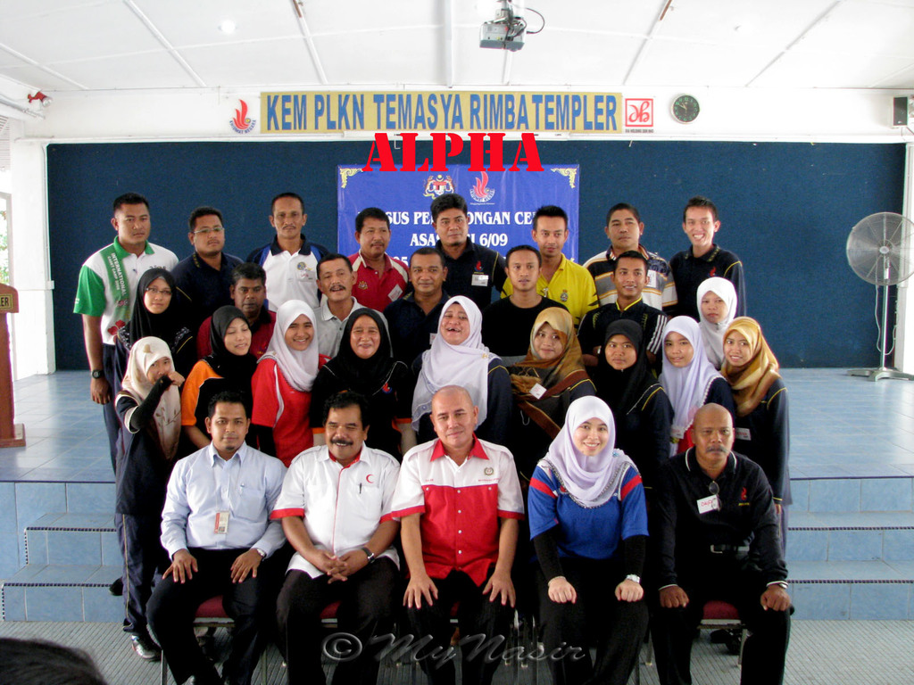 Eine Menschenmenge, die fröhlich vor einer Bühne posiert, mit einem Schild an der Wand im Hintergrund, einem Tischventilator rechts, einem Podium links und einem Projektor oben im Bild.