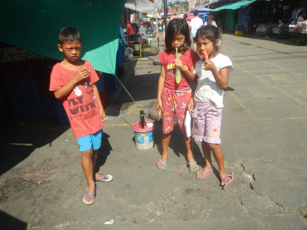 Drei Kinder stehen nebeneinander auf einer Straße, jeder hält ein Eis in der Hand, mit Marktständen, Menschen, Pfählen, Bäumen, Gebäuden und einem klaren blauen Himmel im Hintergrund.