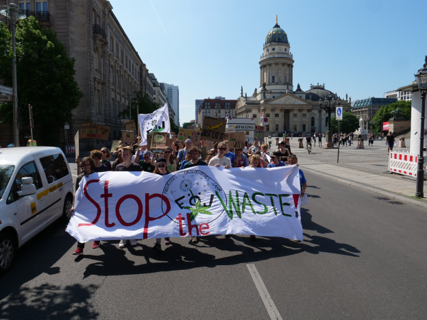 Gruppe von Menschen mit einem "Stop the Waste"-Schild, die eine Straße mit Fahrzeugen, Laternenpfählen, Bäumen, Gebäuden und einem klaren blauen Himmel im Hintergrund überqueren.