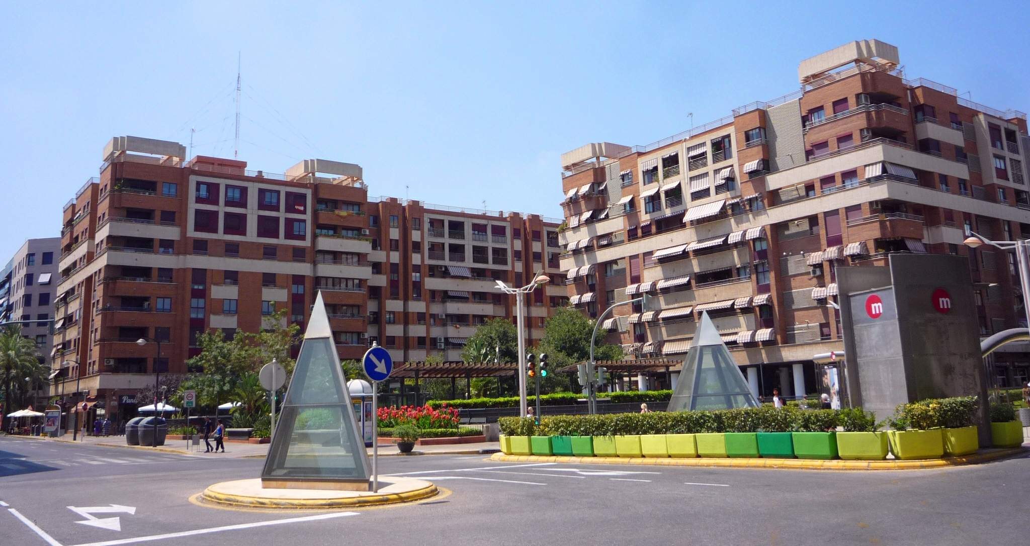 Eine Stadtstraße mit einem zentralen Denkmal, hohen Gebäuden, Straßenmöblierung, Grünfläche, Fußgängern und einem klaren blauen Himmel.