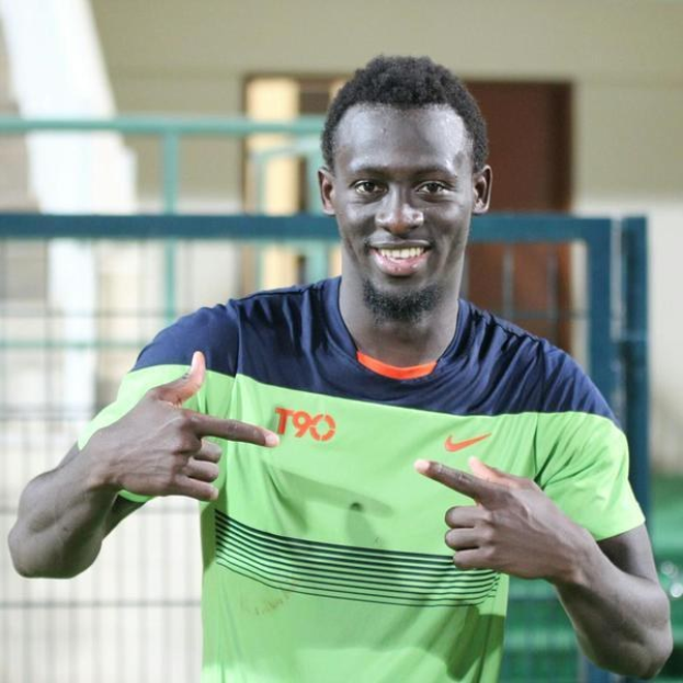 Ein Mann in einem grünen und blauen Hemd lächelt, während er auf die Kamera zeigt, mit einem unscharfen Geländer, Wand und Tür im Hintergrund.