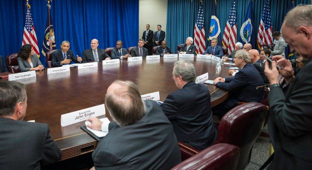 Eine Gruppe von Menschen an einem Tisch mit einigen im Eck stehend, Flaggen vor einem blauen Vorhang