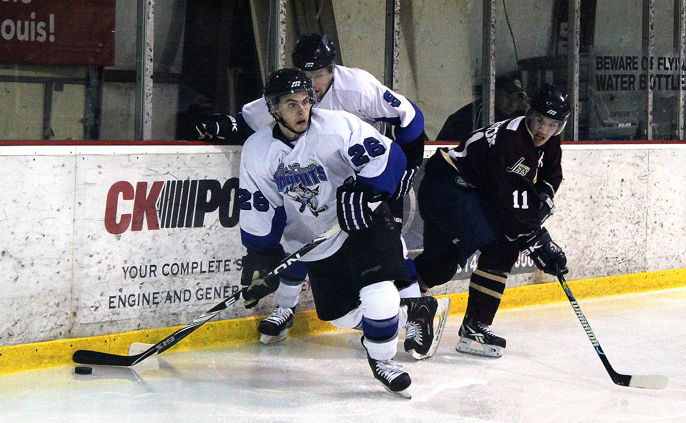 Drei Männer in Hockey-Ausrüstung, jeder mit einem Hockey-Schläger, spielen auf einem Eisstadion mit einer Wand, Text, Pfosten und anderen Gegenständen im Hintergrund.