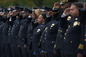 Eine Reihe von Polizeibeamten in Uniform salutiert feierlich vor einem unscharfen Hintergrund aus Bäumen.