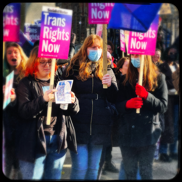Gruppe von Menschen auf einer Straße mit Schildern und Fahnen, darunter eine Frau und ein Mädchen mit 'Trans-Rechte jetzt'-Schildern, mit einem Gebäude im Hintergrund.