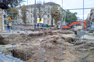 Baustelle mit einem großen Erdloch, das von einem Metallzaun umgeben ist, umgeben von Fahrzeugen, Fußgängern, Verkehrsampeln, Schildern, Bäumen und Gebäuden unter einem klaren blauen Himmel.