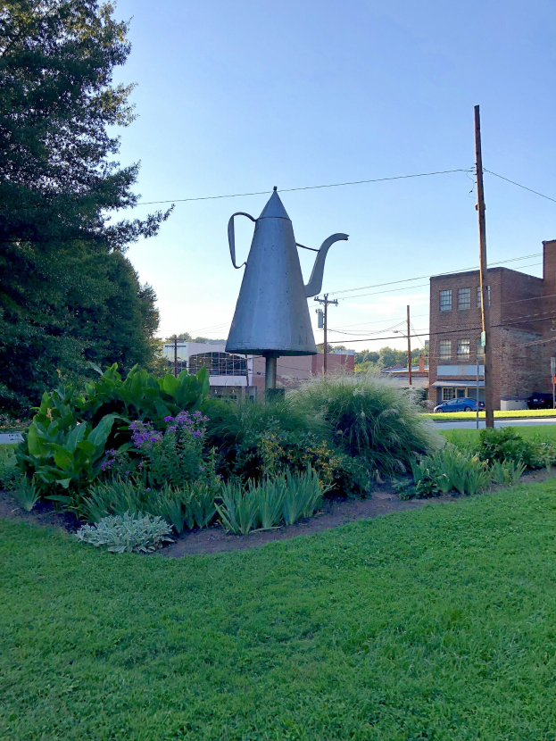 Metallischer Gießkann auf einem grünen Feld mit Pflanzen, Gras, Bäumen, Strommasten, einem Schild, Fahrzeugen, Gebäuden und einem bewölkten Himmel im Hintergrund.
