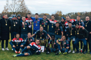 Eine Gruppe von Männern in Medaillen auf einem Fußballfeld mit einem Pokal, umgeben von Bäumen unter einem klaren blauen Himmel.