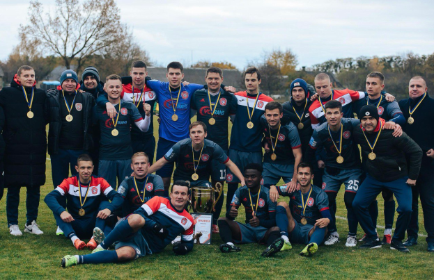 Eine Gruppe von Männern in Medaillen auf einem Fußballfeld mit einem Pokal, umgeben von Bäumen unter einem klaren blauen Himmel.