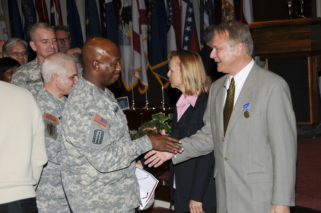 Eine Gruppe von Menschen mit zwei Personen, die sich die Hand schütteln, Flaggen im Hintergrund, ein Podium mit Mikrofon und eine Topfpflanze in der Nähe.