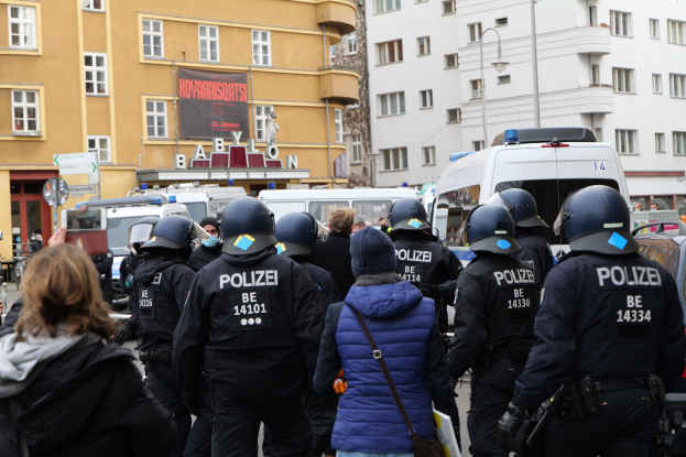 Polizeibeamte in Uniform stehen einer Menge von Helmträgern gegenüber während einer Demonstration in Berlin, mit Fahrzeugen, Gebäuden und einer Kamera im Hintergrund.