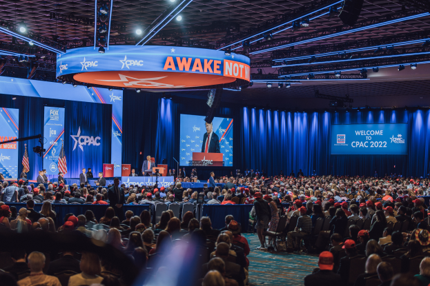 Eine große Menschenmenge, die vor einer Bühne auf einem CPAC 2020-Event sitzt, mit einigen Personen auf dem Podium, blauen Vorhängen und Tafeln mit Text im Hintergrund und einer Decke mit Lampen oben.