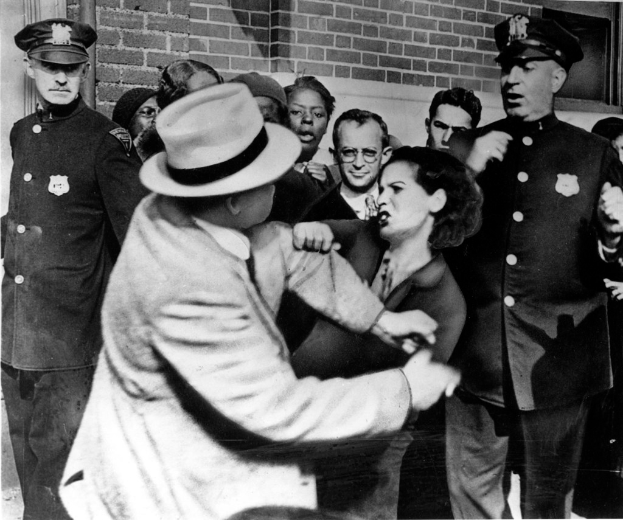 Zwei Personen kämpfen in einem Schwarz-Weiß-Bild, im Hintergrund sind mehrere andere zu sehen, einige tragen Mützen und eine Wand ist im Hintergrund zu erkennen.