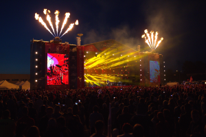 Eine große Menschenmenge auf einem Musikfestival mit einer Bühne in der Mitte, Musiker unter hellen Scheinwerfern und Bannern, Zelte links und ein dunkler Himmel mit Feuerwerk im Hintergrund.