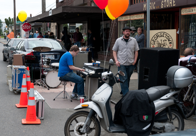 Eine Person, die musikalische Instrumente im Freien spielt, mit Häusern, Bäumen, Menschen, Fahrzeugen und Luftballons im Hintergrund.