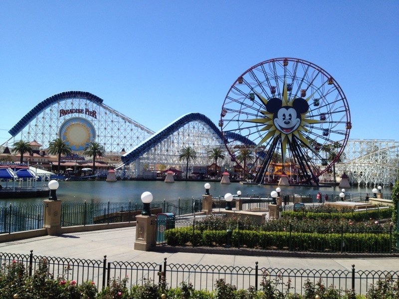 Disneylands California Adventure mit einem Riesenrad und einer Achterbahn, umgeben von Grünflächen und Laternenmasten, mit Wasser im Vordergrund und einem blauen Himmel.