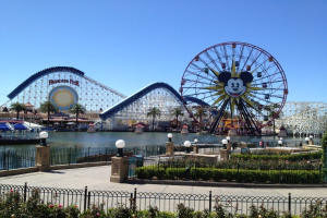 Disneylands California Adventure mit einem Riesenrad und einer Achterbahn, umgeben von Grünflächen und Laternenmasten, mit Wasser im Vordergrund und einem blauen Himmel.