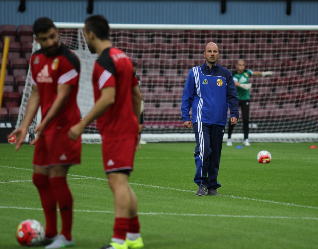 Ein Fußballplatz mit Spielern auf dem Feld, zwei Bälle sichtbar und eine leere Tribüne im Hintergrund.