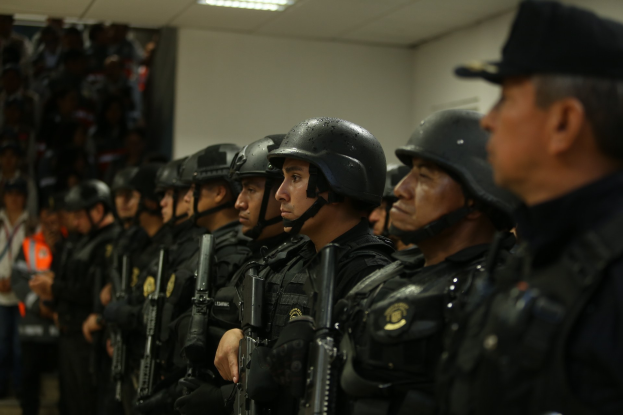 Eine Gruppe von Polizisten in Helmen und Uniformen steht in Formation, jeder hält eine Schusswaffe, mit einer Wand und Deckenbeleuchtung im Hintergrund.
