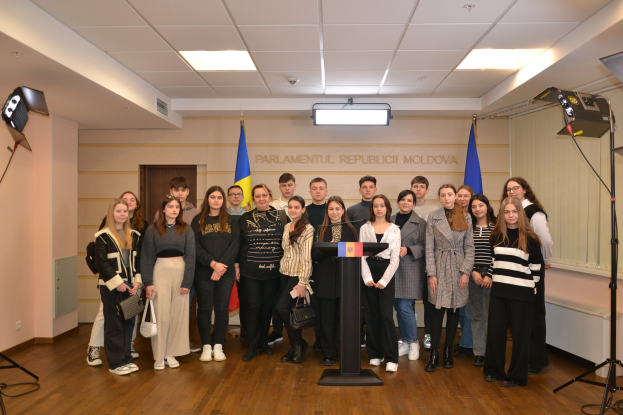 Eine Gruppe von Menschen vor einem Podium mit Mikrofon, umgeben von Fahnen, einer Wand mit dem Text "Moldova Youth Congress 2018", einer T├╝r, einem Dach mit Deckenleuchten und St├Ąnden mit Leuchten.