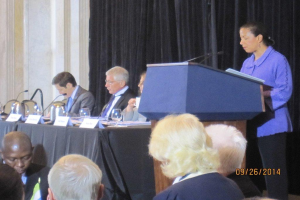 Eine Frau steht an einem Tisch und spricht während einer Konferenz, während drei Männer an einem anderen Tisch mit Gläsern, Büchern und Namensschildern sitzen und einige Menschen im Vordergrund arbeiten und ein Vorhang und eine Wand im Hintergrund zu sehen sind.