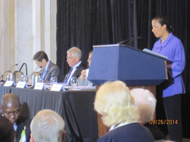 Eine Frau steht an einem Tisch und spricht während einer Konferenz, während drei Männer an einem anderen Tisch mit Gläsern, Büchern und Namensschildern sitzen und einige Menschen im Vordergrund arbeiten und ein Vorhang und eine Wand im Hintergrund zu sehen sind.