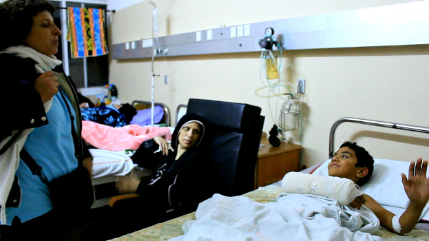 Ein Krankenhauszimmer mit mehreren Betten, auf denen Patienten liegen, Infusionsflaschen auf St├Ąnden zwischen den Betten und eine Frau mit einer Tasche in der N├Ąhe eines Patienten.