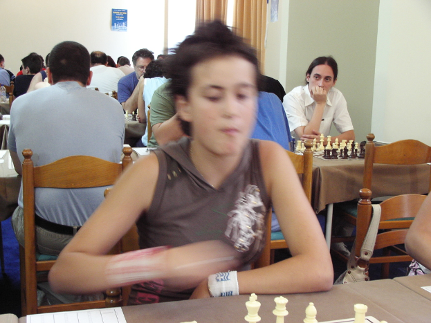 Ein Raum, in dem Menschen an Tischen mit Schachbrettern sitzen und Schach spielen, mit einer Vorhangwand und einem Foto in der rechten Ecke.