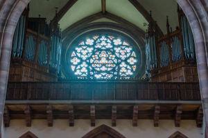 Innenansicht einer Kirche mit einem zentralen Buntglasfenster, flankiert von hölzernen Geländern und Säulen, mit verstreuten Gegenständen und einer Wand unten.