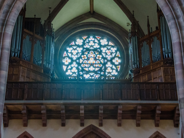 Innenansicht einer Kirche mit einem zentralen Buntglasfenster, flankiert von hölzernen Geländern und Säulen, mit verstreuten Gegenständen und einer Wand unten.
