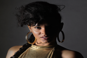 Eine Frau in einem goldenen Kleid und großen Ohrringen steht selbstbewusst vor einer Wand, ihr Haar ist zu einem sleeken Bob gestylt, sie trägt dezentes Make-up mit pinkem Rouge und glänzenden Lippen und schaut direkt in die Kamera mit einem entschlossenen Ausdruck.