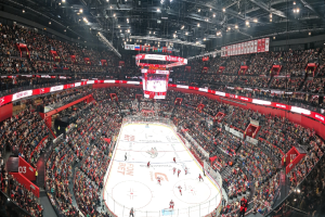 Eine große, hell erleuchtete Arena voller jubelnder Zuschauer, die ein Eishockeyspiel verfolgen, mit einem zentralen Bildschirm über der Eisfläche.