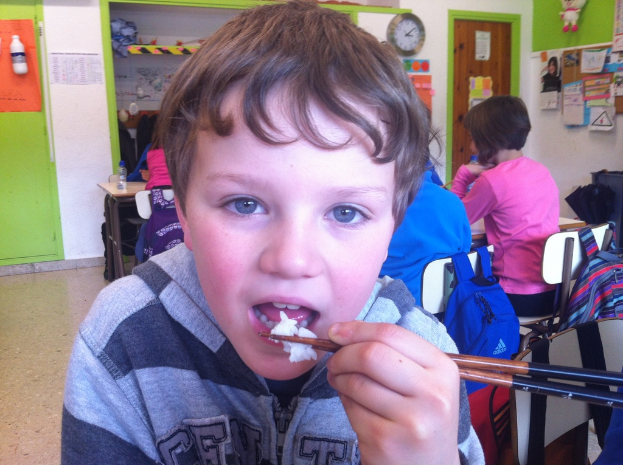 Ein Junge in einer grauen und schwarzen Jacke isst mit Stäbchen in einem Klassenzimmer, mit anderen Kindern im Hintergrund, einer grün-weißen Wand, Lehrplakaten an einer weißen Wand, einer braunen Tür und einer Wanduhr.
