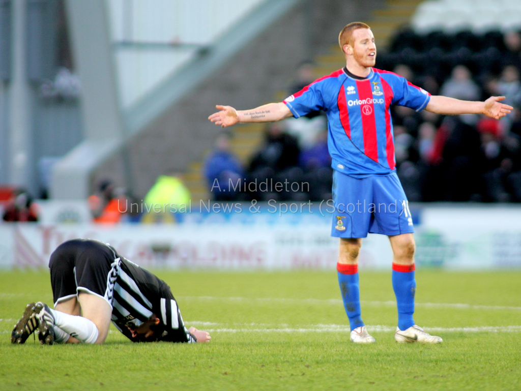 Ein Stadion mit einer Person, die steht, und einer anderen, die kniet, auf dem Gras im Vordergrund, und einer Gruppe von Menschen mit Schildern im Hintergrund.