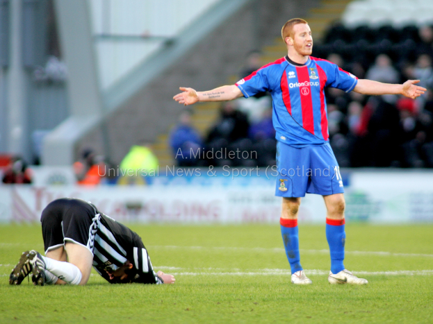 Ein Stadion mit einer Person, die steht, und einer anderen, die kniet, auf dem Gras im Vordergrund, und einer Gruppe von Menschen mit Schildern im Hintergrund.