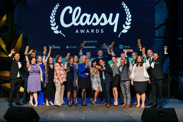 Gruppe von Menschen auf einer Bühne mit Auszeichnungen, mit Lautsprechern darunter und einem Bildschirm mit der Aufschrift "Classy Awards 2019" im Hintergrund.