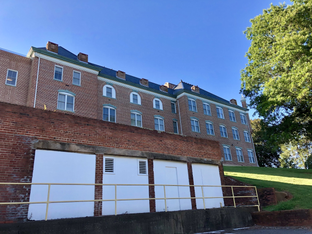 Großes Backsteingebäude mit zahlreichen Fenstern, umgeben von einem Metallzaun, Weg, Gras, Bäumen und einem bewölkten Himmel, auf dem Campus der University of Virginia.