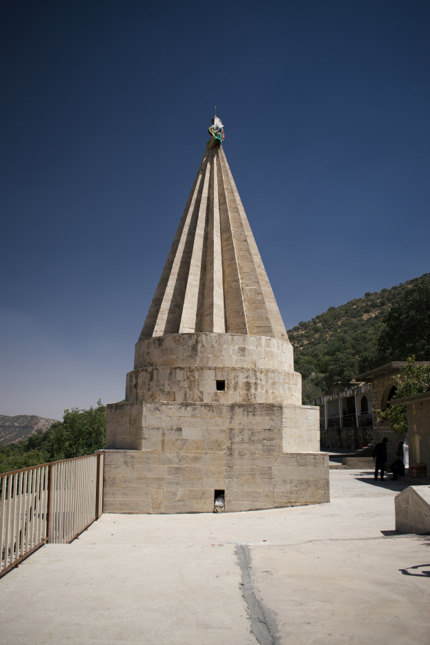 Ein Denkmal in einem Park mit einem Geländer auf der linken Seite, umgeben von ein paar Menschen, mit Bäumen, Hügeln und einem klaren blauen Himmel im Hintergrund.